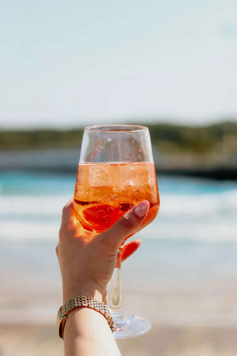 A hand holding an Aperol Spritz on the beach